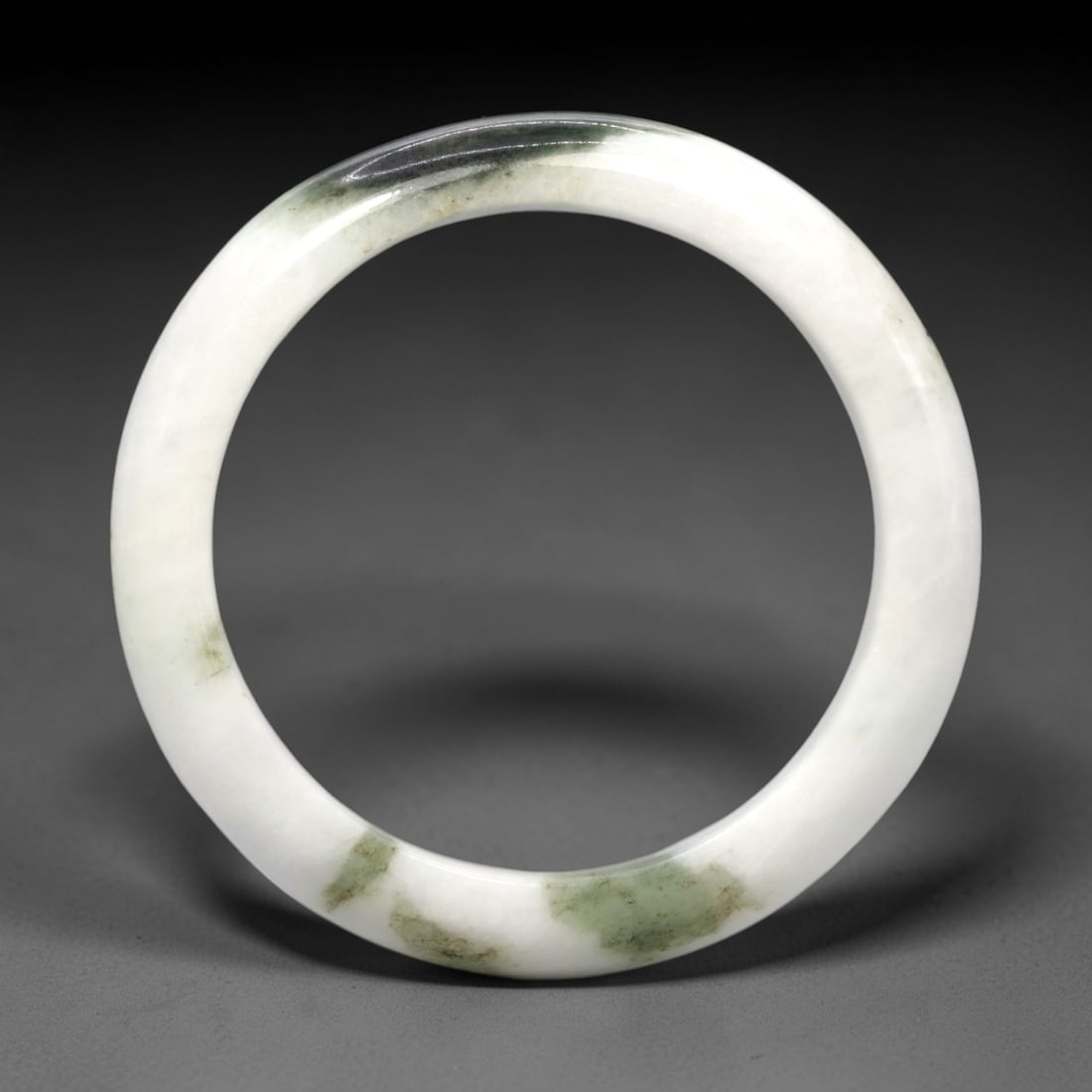 A Burmese Jadeite Jade Bangle, 20th Century, 16FS: A Burmese jadeite jade bangle, with a pale white matrix and scattered dark green mineral inclusions. The bangle is of circular form, with a smooth, polished surface revealing the internal crystalline