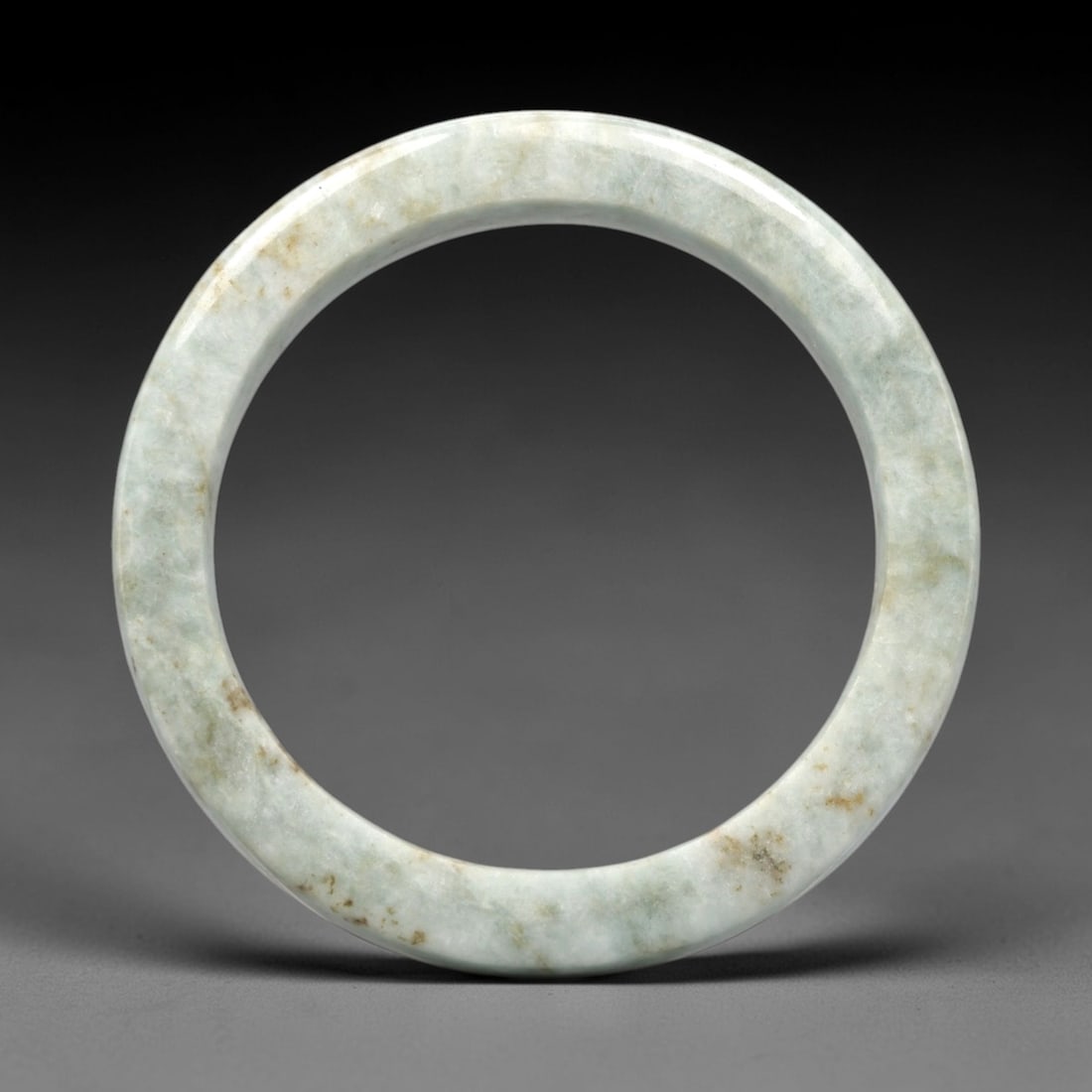 A Burmese Jadeite Jade Bangle, 20th Century, 20FS: A Burmese jadeite jade bangle, with a pale greenish-white matrix and scattered russet-brown mineral inclusions. The bangle is of circular form, with a smooth, polished surface revealing the internal c