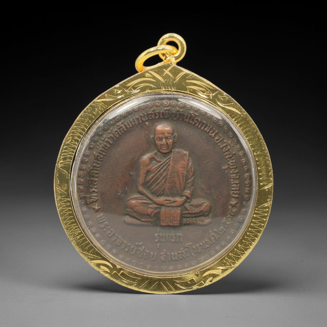 A Thai Copper Alloy Buddhist Amulet in Gold Frame, Late 20th Century, 99FP: An oval-shaped copper alloy amulet, measuring 4.5 x 3.8 cm, set within an engraved gold frame with a suspension loop. The obverse depicts a seated Buddhist monk in meditative mudra, encircled by a con
