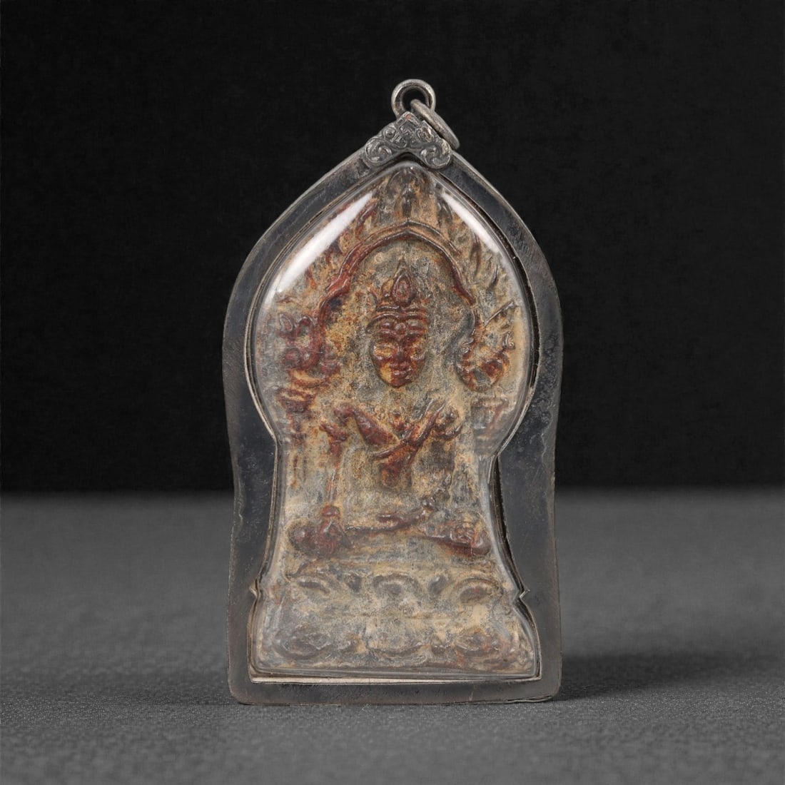 A Thai Seated Deity Amulet with Silver Frame, 20th Century, 34FP: The amulet is of shield-shaped form, featuring a molded seated deity figure on the obverse, rendered in a dark patinated material against a light beige ground. The deity is depicted with multiple arms