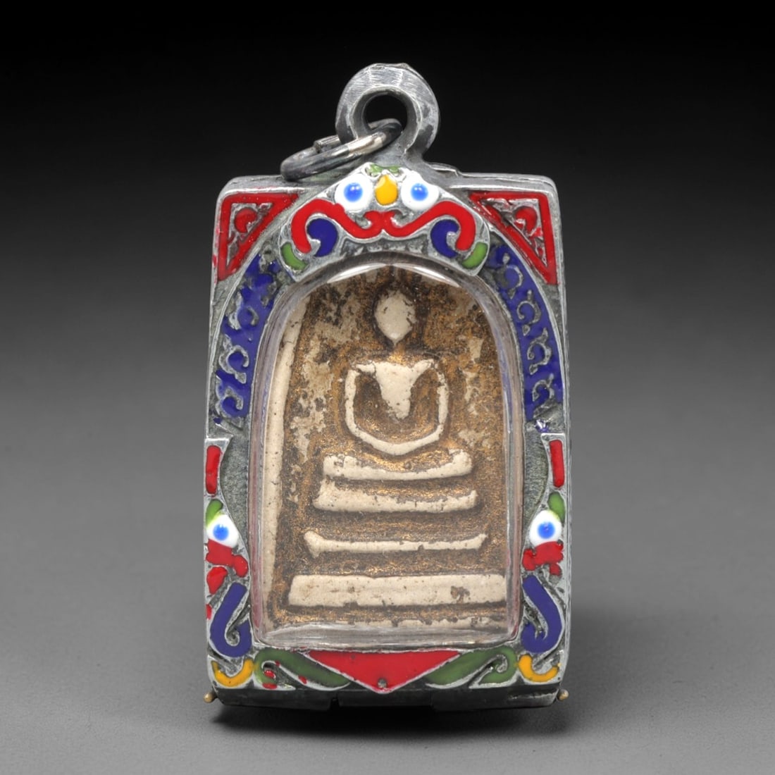 A Thai Phra Somdet Clay Amulet in a Polychrome Enamel Silver-Tone Case, 20th Century, 14ZK: This revered Phra Somdet amulet is fashioned from sacred beige clay and housed in an ornate, vintage silver-tone metal case with a prominent suspension loop at the apex. The obverse of the case featur