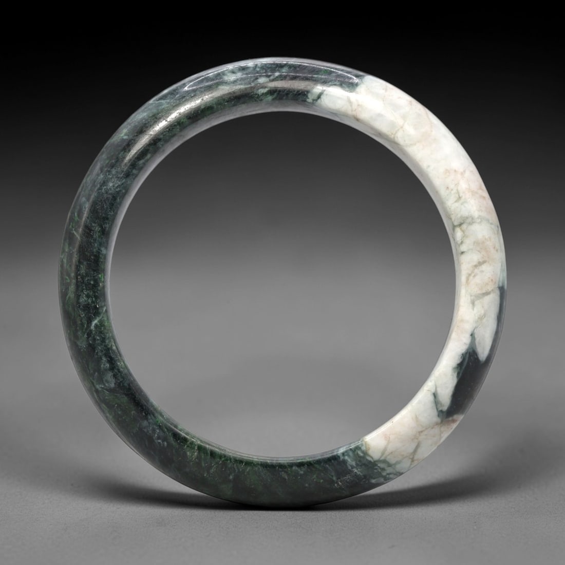 A Burmese Jadeite Jade Bangle, 20th Century, 10FS: A Burmese jadeite jade bangle, with a dark green matrix and prominent white mineral inclusions. The bangle is of circular form, with a smooth, polished surface revealing the internal crystalline struc