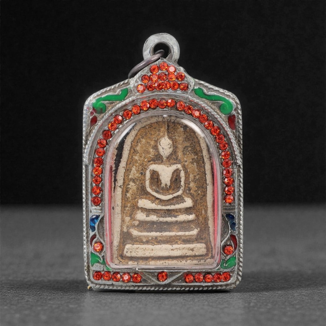 A Thai Buddha Amulet with Silvered Frame, 20th Century, 40FP: The amulet is of rectangular form with a pointed upper edge. The obverse features a molded seated Buddha figure in meditative pose (dhyana mudra), rendered in a light-colored material against a tan gr