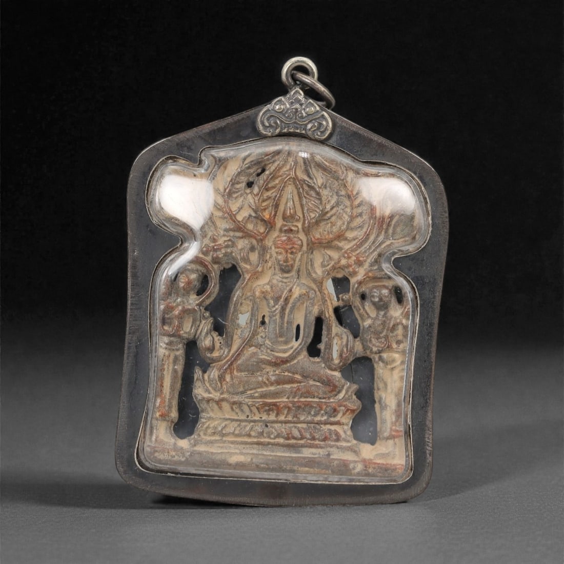 A Thai Seated Buddha Amulet with Metal Frame, 20th Century, 32FP: The amulet is of shield-shaped form, featuring a molded seated Buddha figure on the obverse, rendered in a dark patinated material against a light beige ground. The Buddha is depicted seated on a lotu