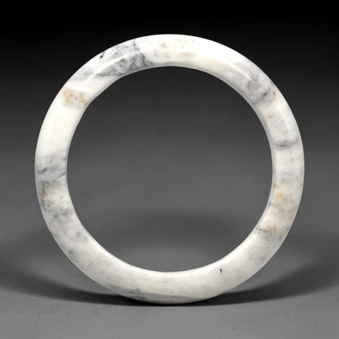 A Burmese Jadeite Jade Bangle, 20th Century, 19FS: A Burmese jadeite jade bangle, with a pale white matrix and scattered dark grey and russet-brown mineral inclusions. The bangle is of circular form, with a smooth, polished surface revealing the inter