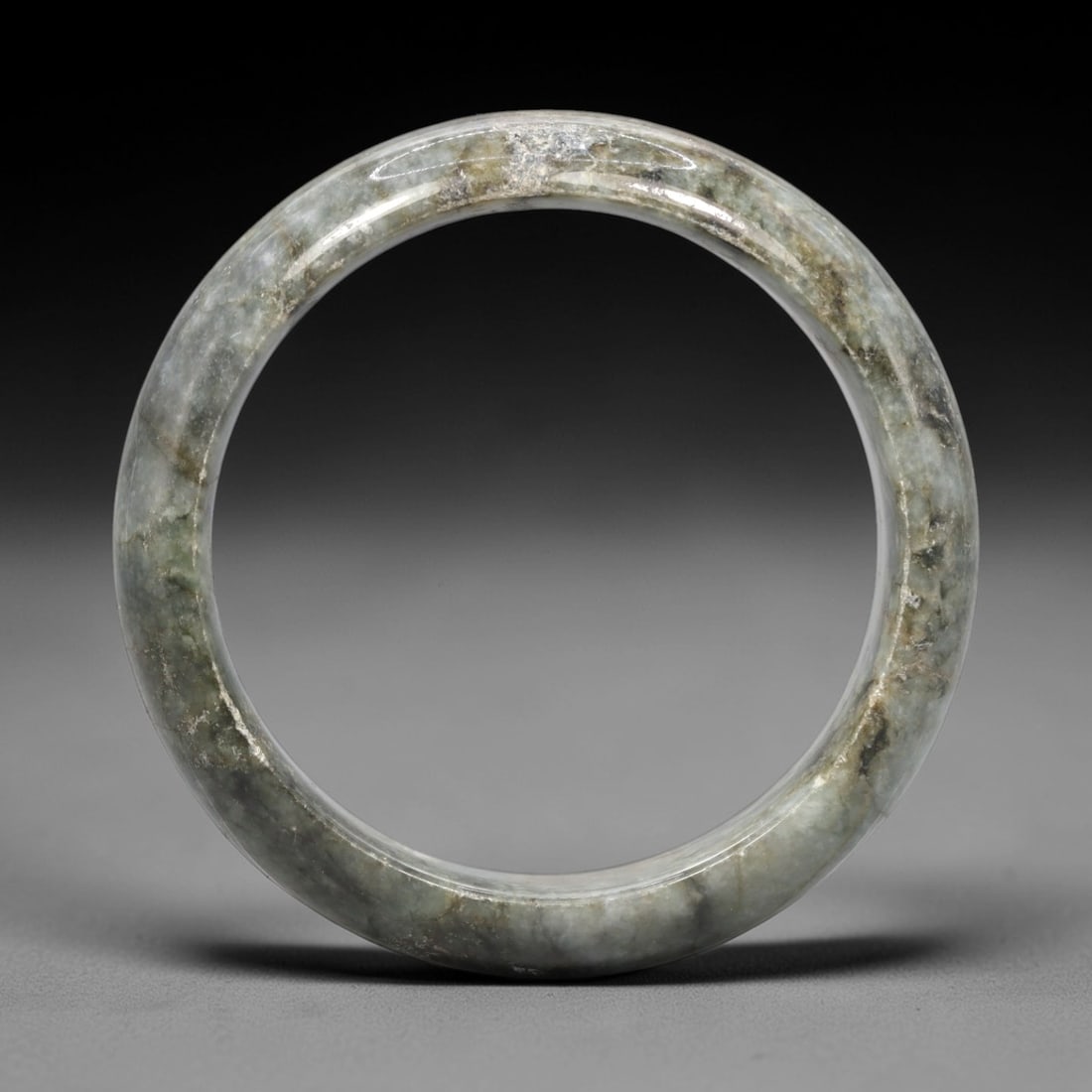 A Burmese Jadeite Jade Bangle, 20th Century, 4FS: A Burmese jadeite jade bangle, with a pale greyish-green matrix and prominent darker green mineral inclusions. The bangle is of circular form, with a smooth, polished surface revealing the internal cr