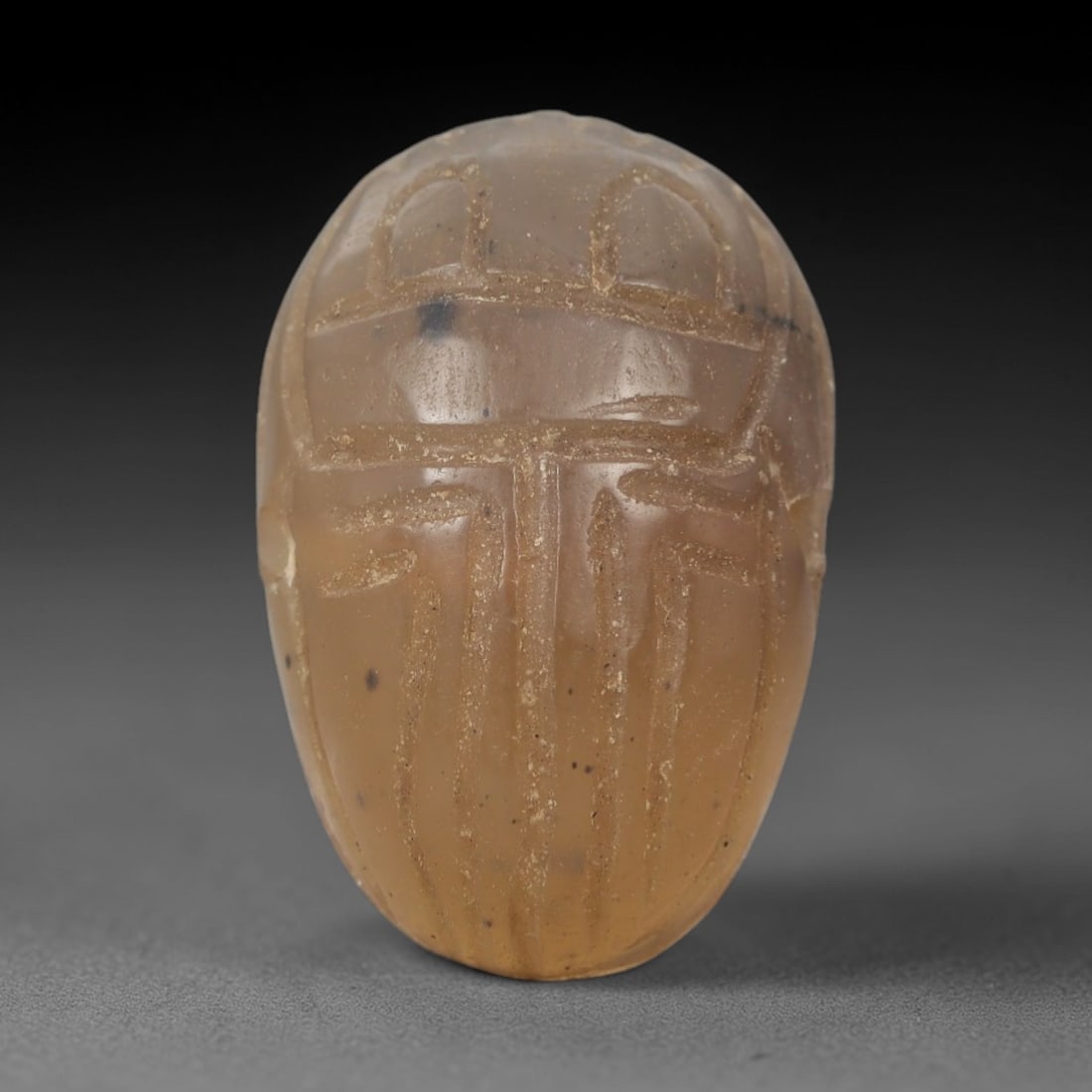 Ancient Chalcedony Scarab Amulet - Egyptian Figurine, 66AJ: Carved from translucent pale chalcedony with subtle amber undertones and natural dark mineral inclusions, this amulet is formed as a stylized scarab beetle with deeply incised linear detailing to defi