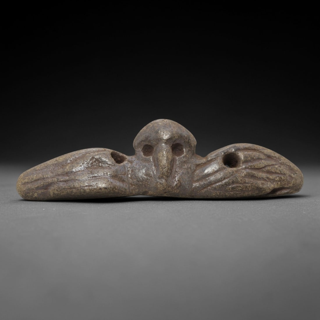 Ancient Steatite Bird Amulet - Near Eastern Ritual Pendant, 88FW: Carved from a dense, speckled steatite stone, this amulet depicts a stylized avian form in a horizontal, outstretched configuration, featuring a central head with recessed eye sockets and a defined be