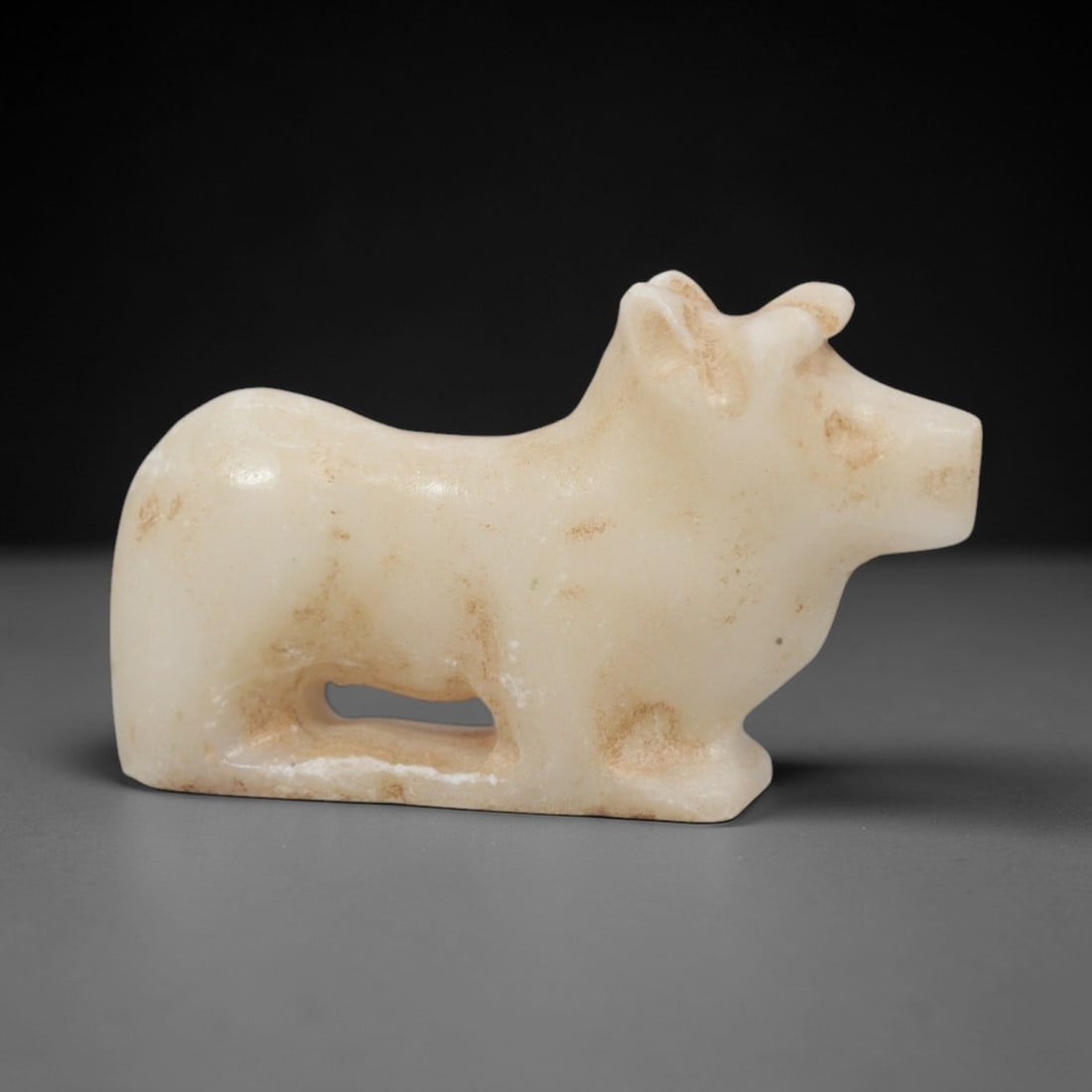 Ancient Marble Bovid - Near Eastern Figurine, 94XR: Crafted from pale marble, this small figurine depicts a recumbent bovid in a stylized, compact form. The robust body rests on a solid base, with a rectangular suspension hole carved beneath the torso,