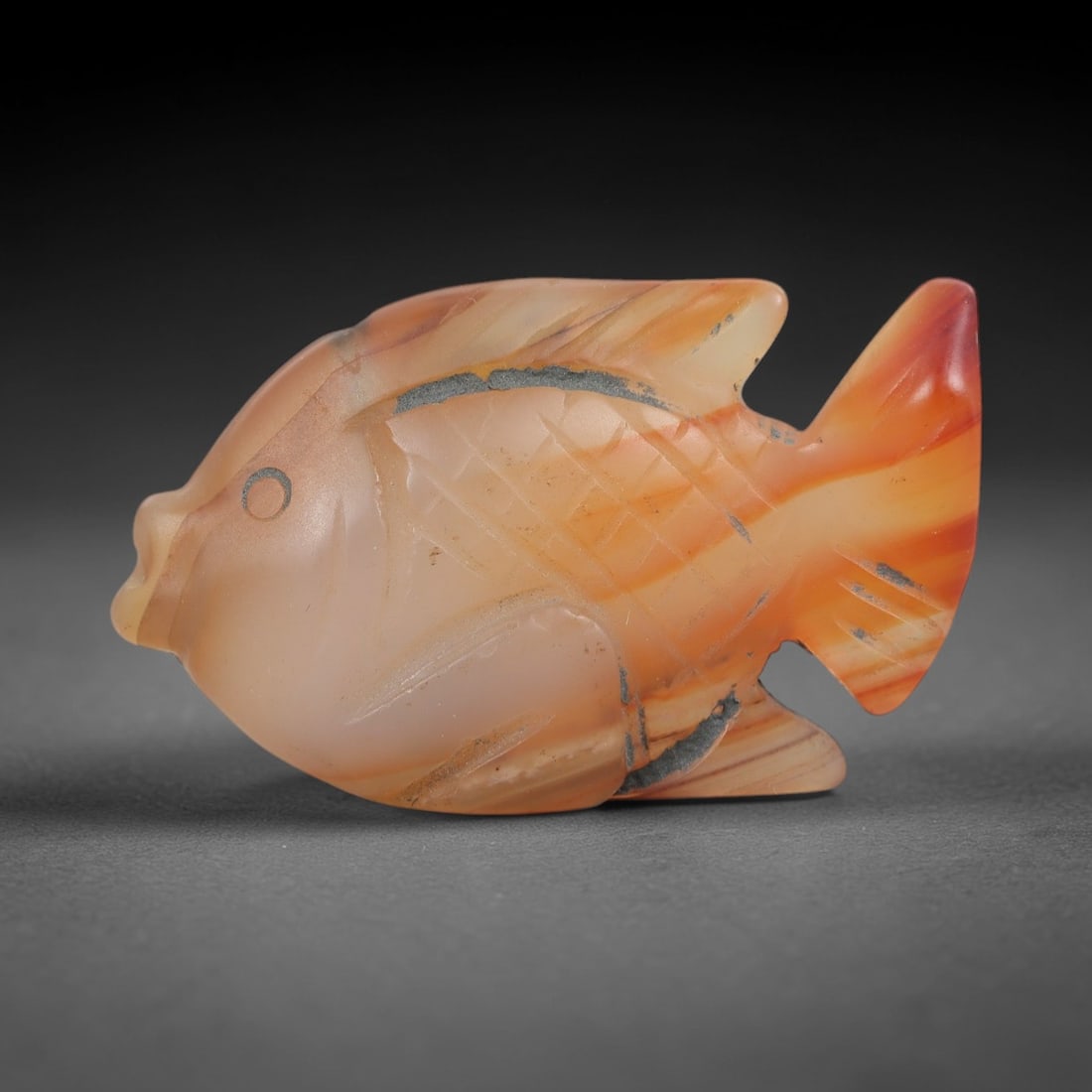 Ancient Agate Fish Figurine - Egyptian Carving, 41AJ: Carved from banded orange-and-peach agate, this piece is formed as a detailed fish with incised cross-hatching across the body to denote scales, defined dorsal, pectoral, and tail fins, and a stylized