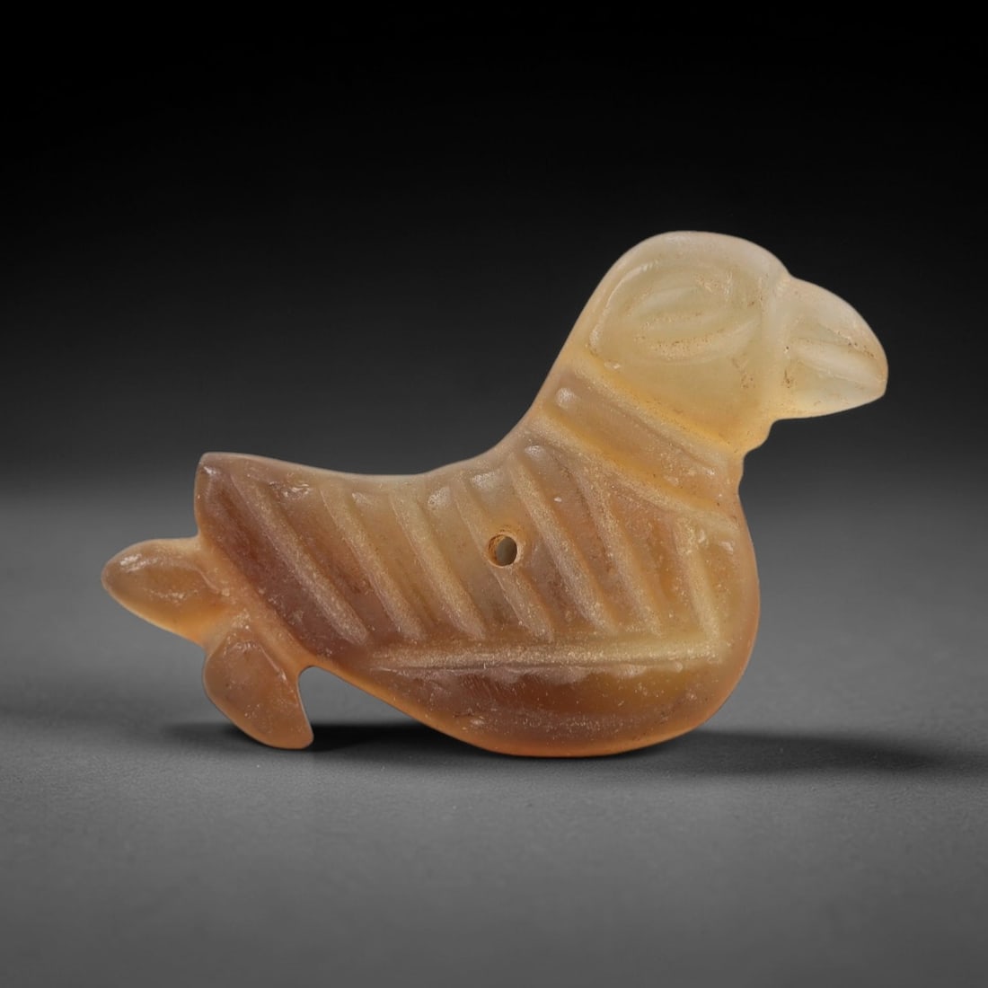Ancient Chalcedony Bird Amulet - Egyptian Figurine, 57AJ: Carved from translucent warm-toned chalcedony, this amulet is formed as a stylized bird with a prominent beak, incised linear detailing across the body to denote feather patterns, and a distinct fanne