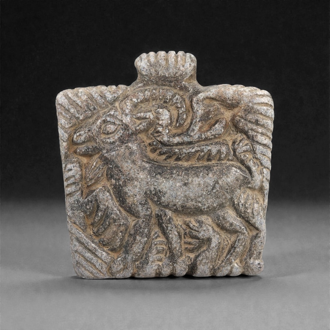 A Syro-Anatolian Schist Zoomorphic Amulet, Early 1st Millennium BCE, 54LJ: Carved from dark grey schist, the amulet presents a horned quadruped in profile, shown walking with the limbs extended and the head slightly raised. A large, backward-curving horn is rendered in low r