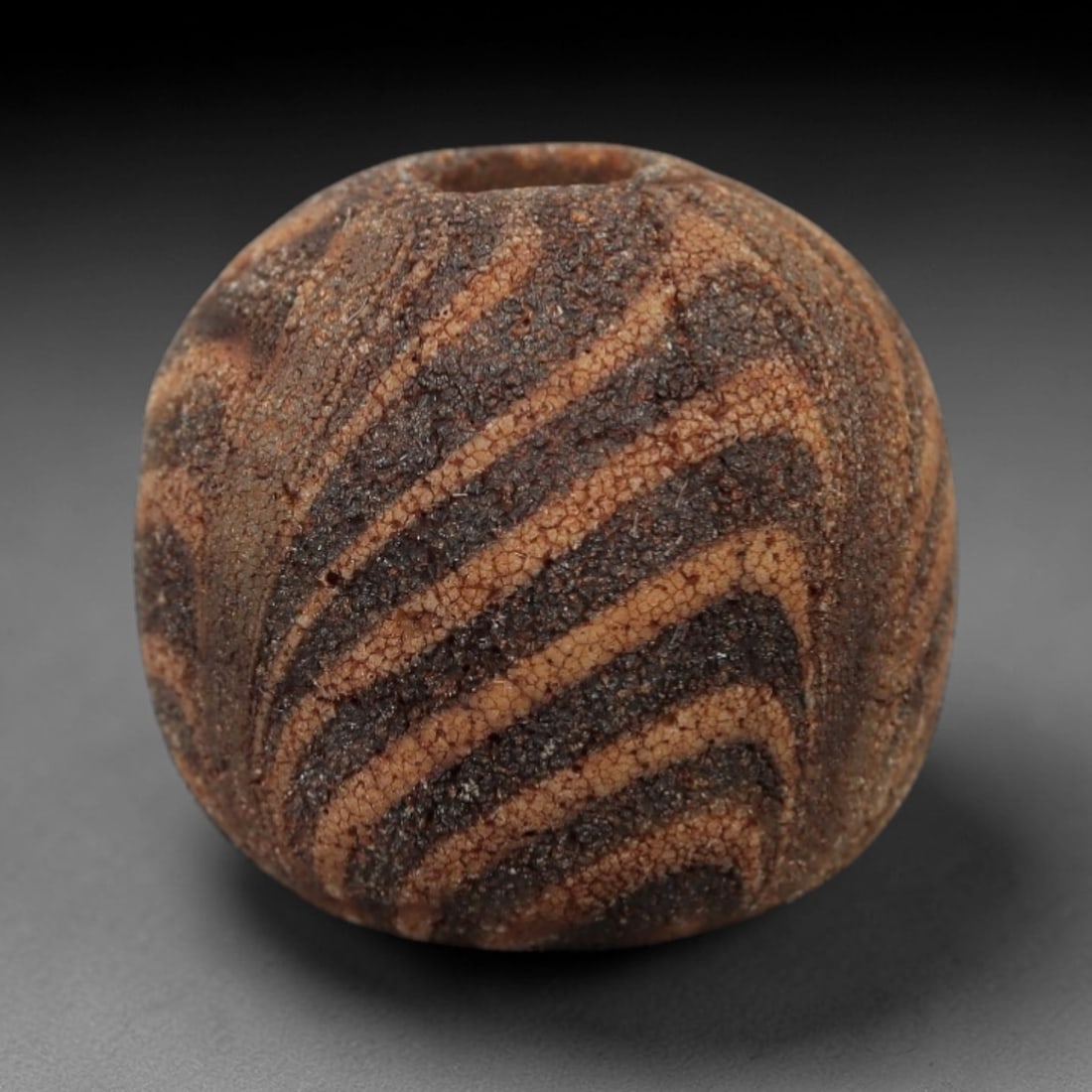 Ancient Islamic Glass Bead - Chevron Stripe Amulet Bead, 55FW: This spherical bead is crafted from porous, earth-toned glass, featuring bold diagonal chevron stripes in warm tan and deep brown-characteristic of medieval Islamic glassworking traditions. A central