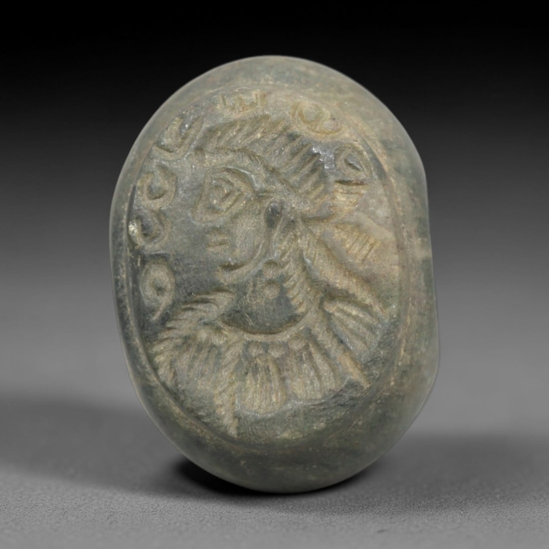 Ancient Stone Hemispherical Stamp Seal - Near Eastern Profile Figure, 44ZP: Crafted from a fine-grained, greenish stone, this hemispherical stamp seal bears a deeply incised profile depiction of a Near Eastern figure, adorned with a stylized headdress and elaborate neck ornam