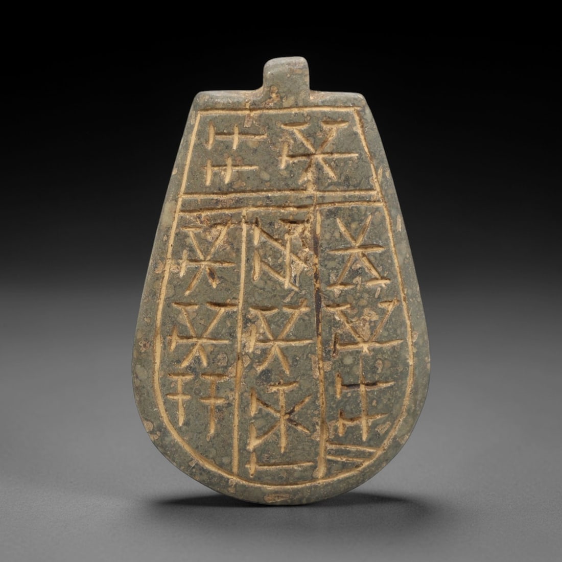 Ancient Near Eastern Stone Inscribed Amulet - Pendant, 35ZR: This artifact is a carved stone inscribed amulet pendant, crafted from a fine-grained, grey-green stone with a matte surface and earthy patina filling the incised linear inscriptions. The teardrop-sha