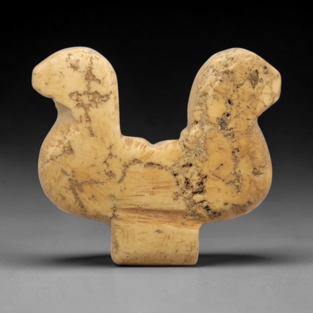 Ancient Ivory Double-Headed Animal - Near Eastern Figurine, 51ZW: This object is carved from pale ivory, depicting a double-headed animal with two stylized heads facing opposite directions, each with simplified facial contours. The body is geometrically simplified, 