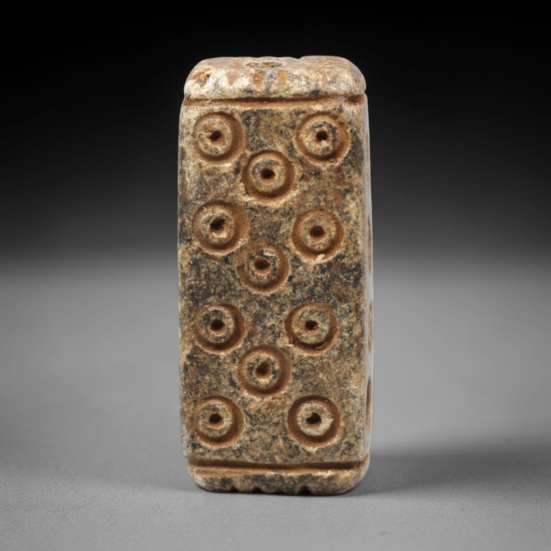 Ancient Steatite Amulet Bead - Near Eastern Concentric Circles, 54XV: Carved from dense steatite, this ancient amulet bead features an elongated rectangular form with a vertical suspension hole at the top, designed for wearable use. Its face is decorated with a symmetri