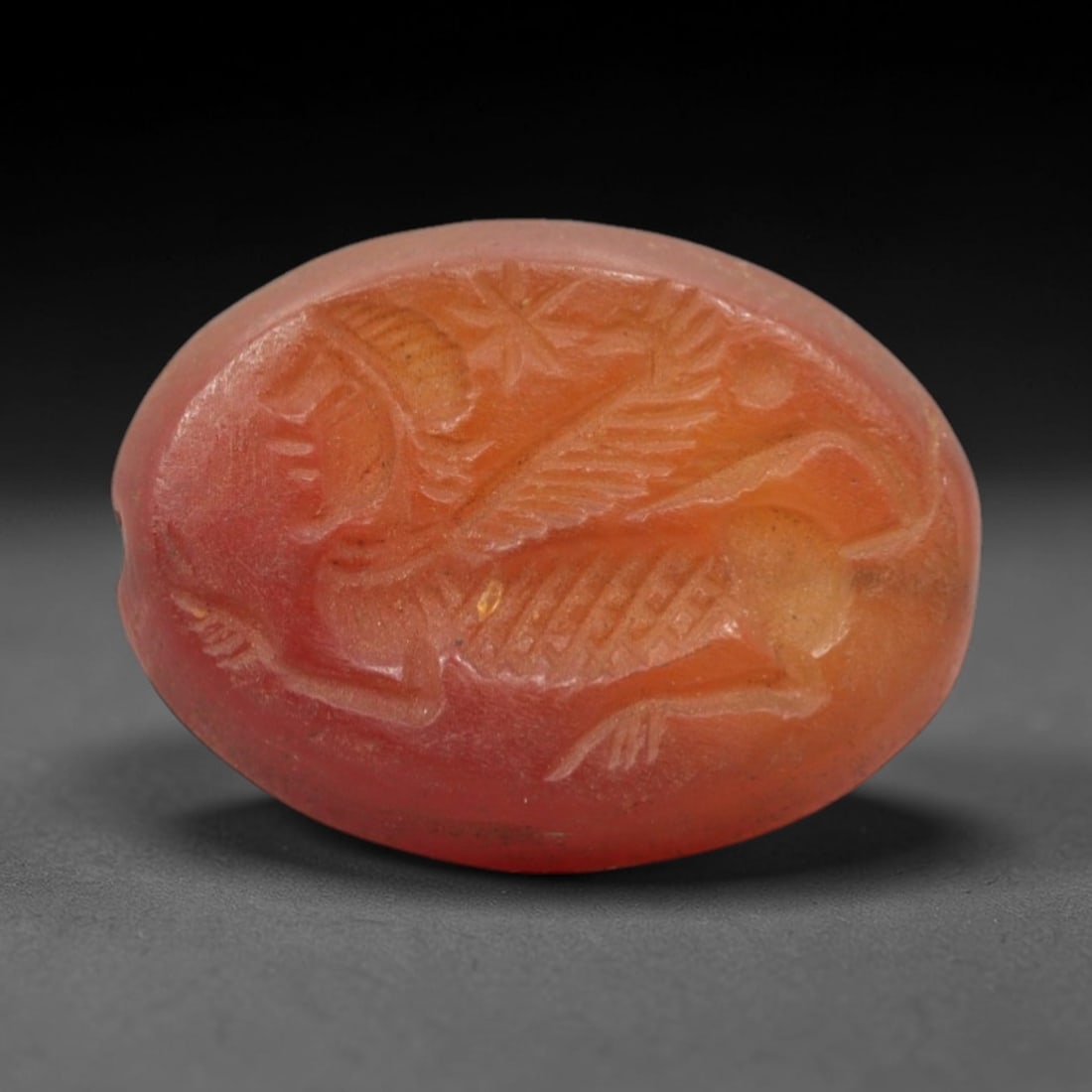 Ancient Carnelian Intaglio - Winged Sphinx/Harpp Figure Seal, 31XV: Crafted from polished warm-toned carnelian, this oval intaglio seal depicts a striding winged hybrid creature-likely a sphinx or harpy-with a human head, feathered wings, and a leonine body. The figur