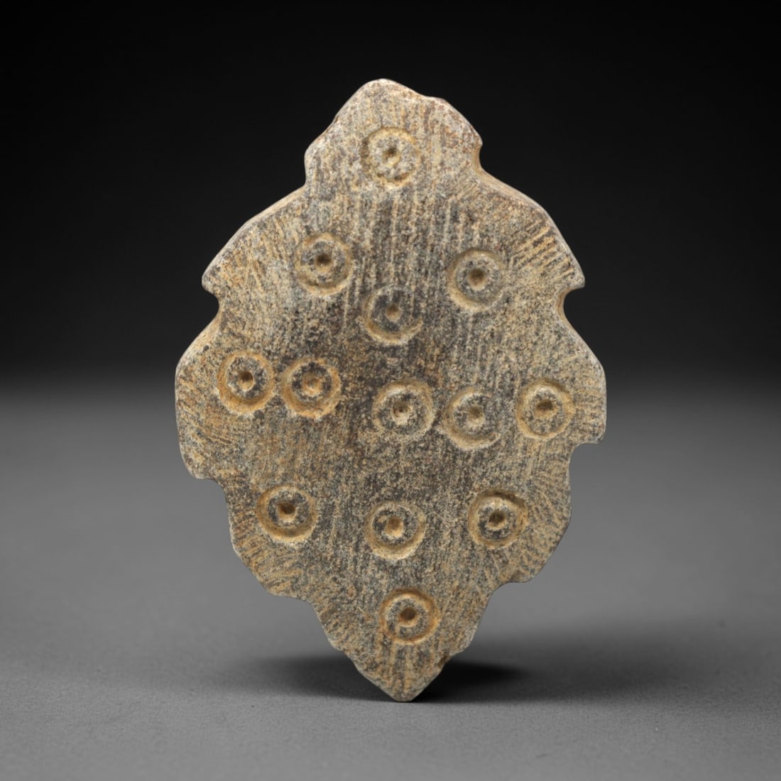 Ancient Steatite Amulet - Near Eastern Concentric Circles, 56XV: Carved from dense steatite, this ancient amulet exhibits a distinctive foliate or petal-like form with a scalloped, undulating edge, creating a symmetrical and elegant silhouette. Its surface is adorn