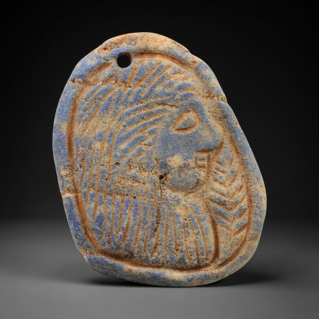 Ancient Lapis Lazuli Plaque - Near Eastern Amulet, 9AJ: Carved from lapis lazuli stone, this ancient Near Eastern plaque features a stylized human head with detailed facial features and a crown-like headdress. The carving exhibits fine craftsmanship typica