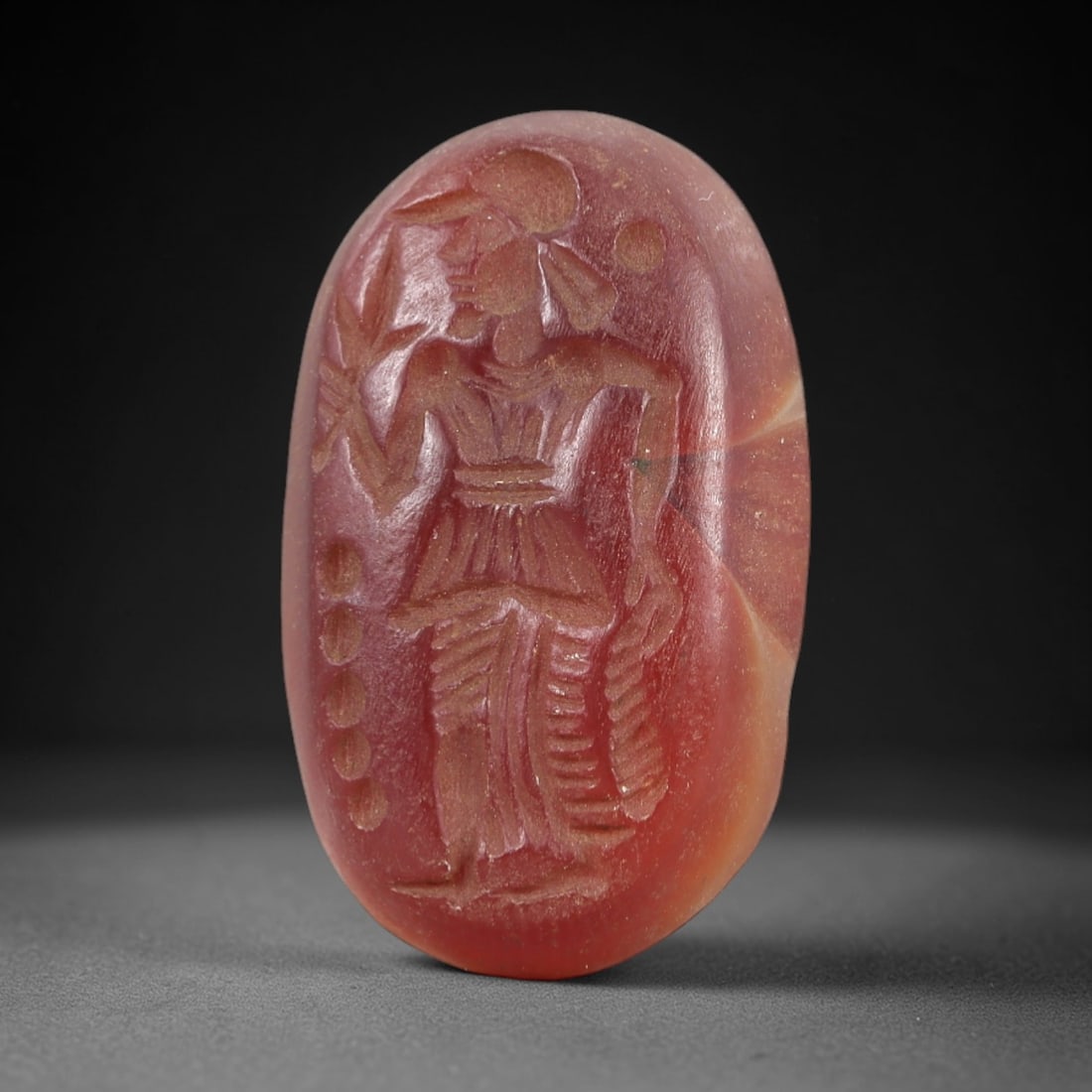 Ancient Carnelian Intaglio - Near Eastern Figural Amulet, 11AL: This oval intaglio is carved from polished carnelian, featuring a standing human figure in relief, clad in pleated garments and a crested head covering, holding a leafy branch in one hand and a coiled