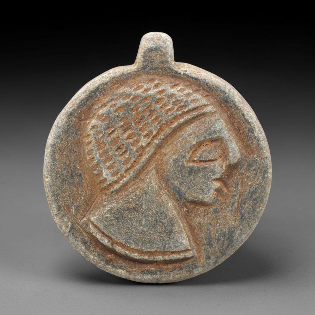 Ancient Stone Anthropomorphic Amulet - Near Eastern Profile Figurine, 23ZR: This artifact is a circular stone anthropomorphic amulet, executed in the stylized profile tradition of the ancient Near East. The face depicts a human figure in right profile, with a textured, dotted