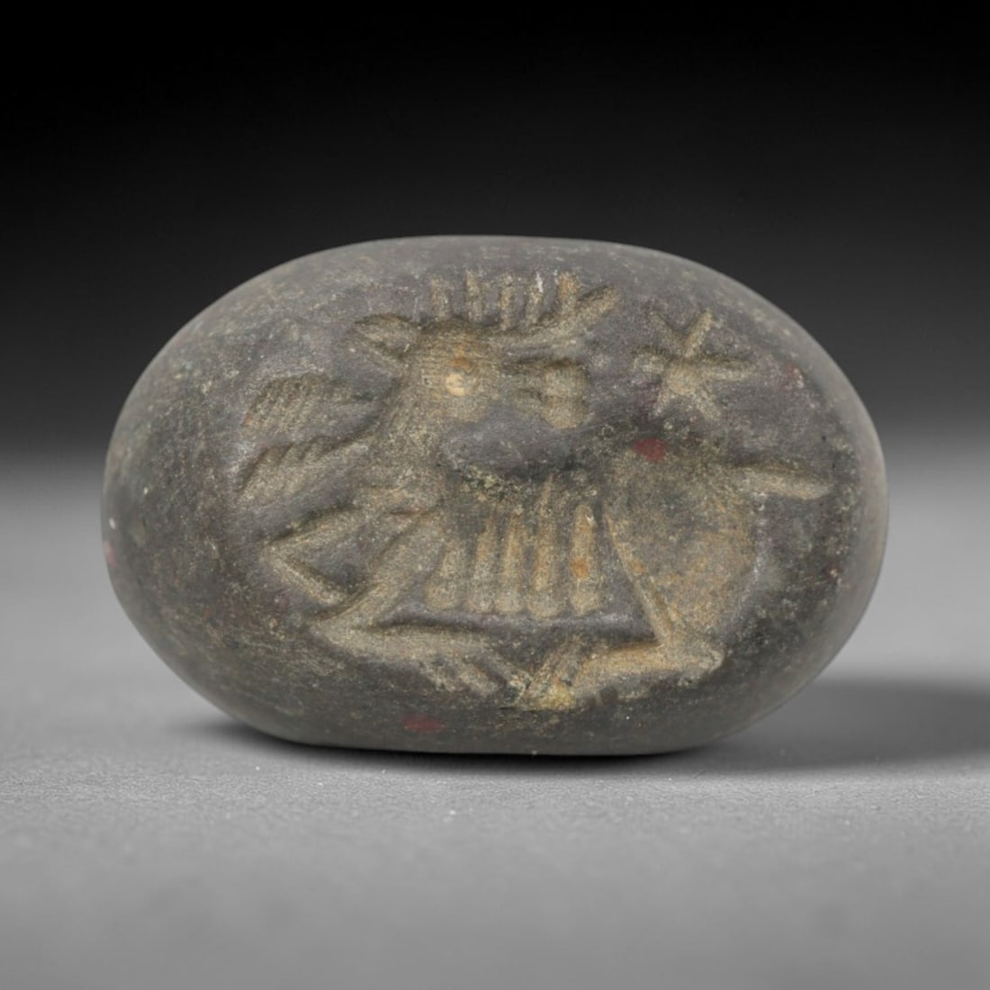 Ancient Stone Hemispherical Stamp Seal - Near Eastern Ibex Star Figure, 79ZS: Crafted from dense, dark gray stone, this hemispherical stamp seal bears incised carving of a leaping ibex, with prominent curved horns, segmented torso, and dynamic linear limb motifs, alongside a st