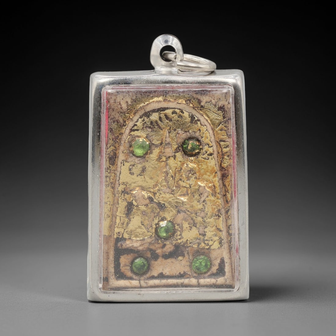 A Thai Terracotta Amulet in Metal Case, 20th Century, 62ZH: The amulet is formed from terracotta, with a light beige matrix overlaid with a gold-toned layer on the obverse. The obverse depicts a seated figure within a recessed arched frame, with five green fac