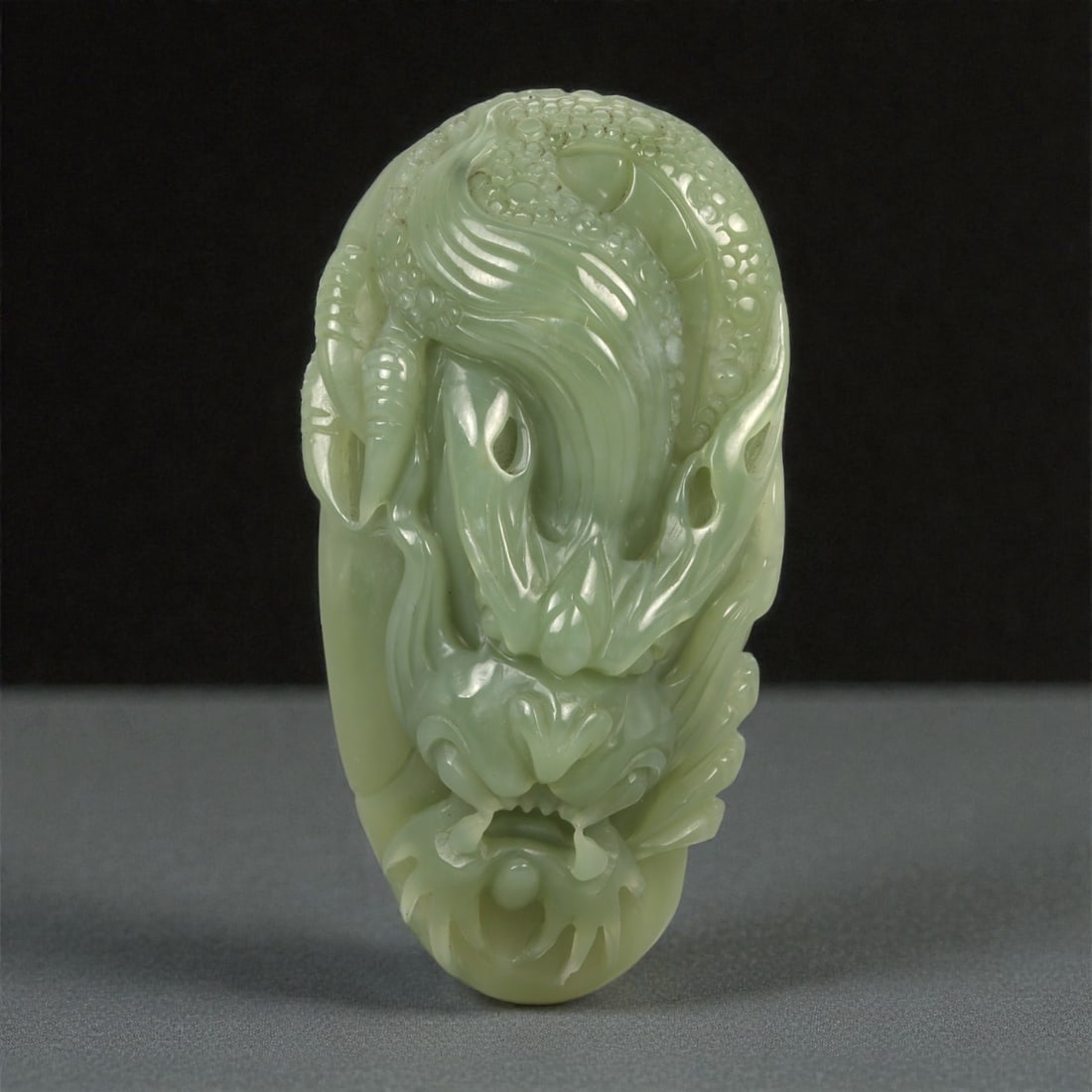 A Chinese Jade Mythical Beast Carving, 20th Century, 42YA: Carved from pale celadon jade with soft translucency and subtle tonal variation, the object is modeled as a vertically oriented mythical beast head rendered in high relief. The composition is dominate