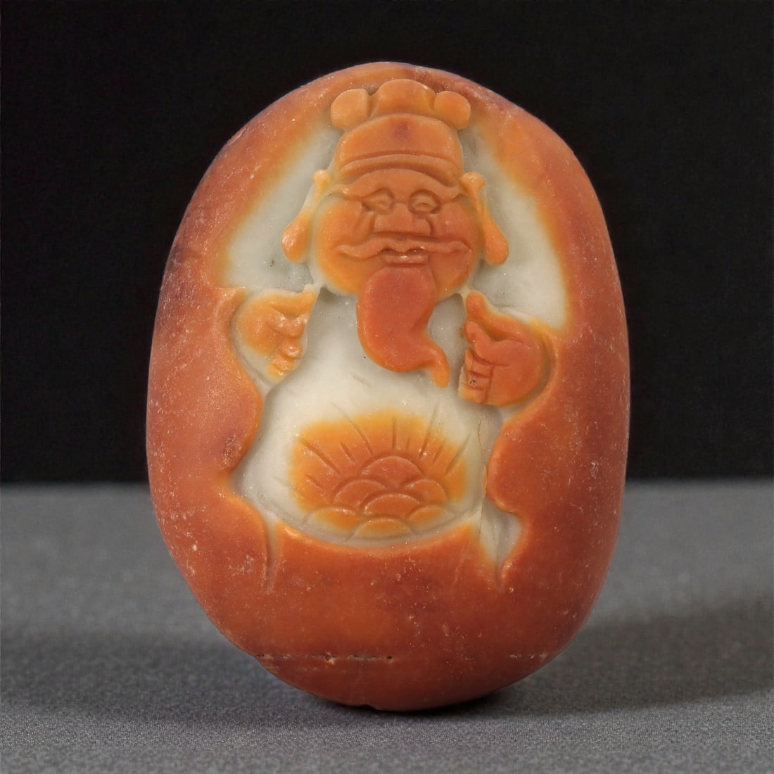 A Chinese Hardstone Figure Plaque, 20th Century, 74FC: Carved from reddish-orange hardstone with areas of translucent white matrix, the object is worked as an oval plaque featuring a standing male figure in relief. The figure is shown frontal, wearing a s