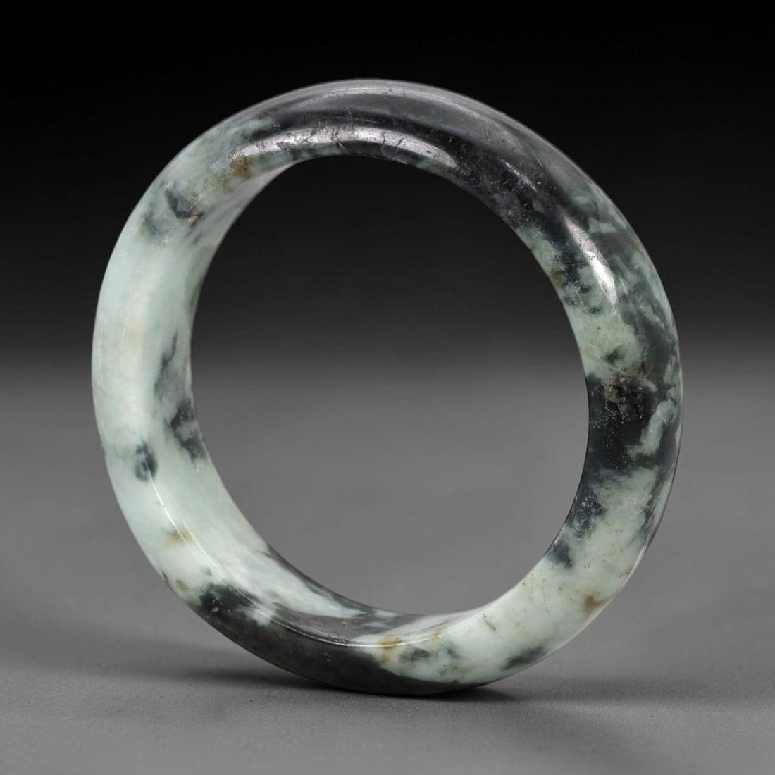 A Chinese Jadeite Jade Bangle Bracelet, 20th Century, 96ZK: Carved from jadeite jade with a striking variegated composition of pale milky-green and deep blackish-green mottling, the bangle is formed as a solid circular band with a flattened rounded cross-secti