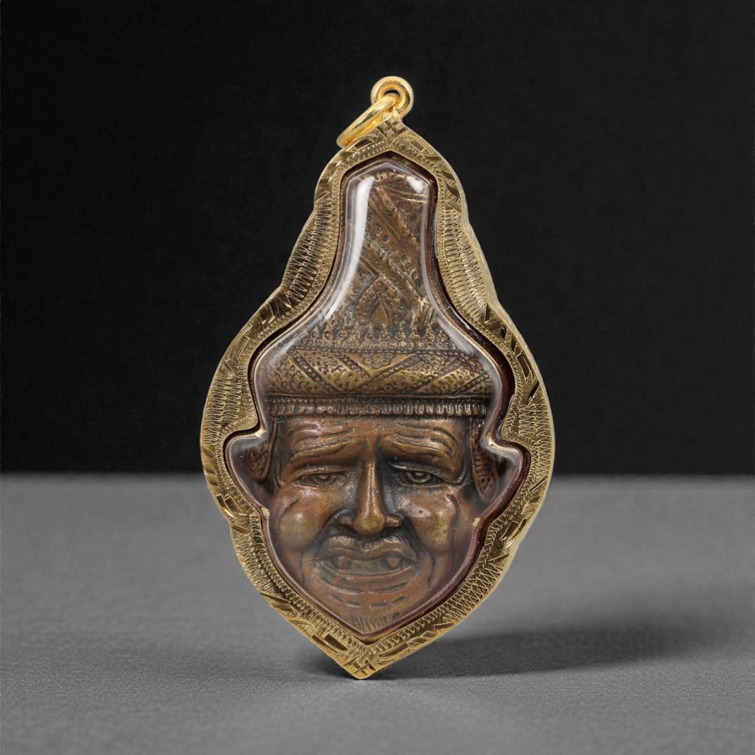 A Thai Copper Amulet in Gilt Metal Frame, 20th Century, 60FN: The amulet is housed in a teardrop-shaped gilt metal frame with incised scrollwork borders and a suspension loop at the apex. The obverse features a copper high-relief carving of a human face with exa
