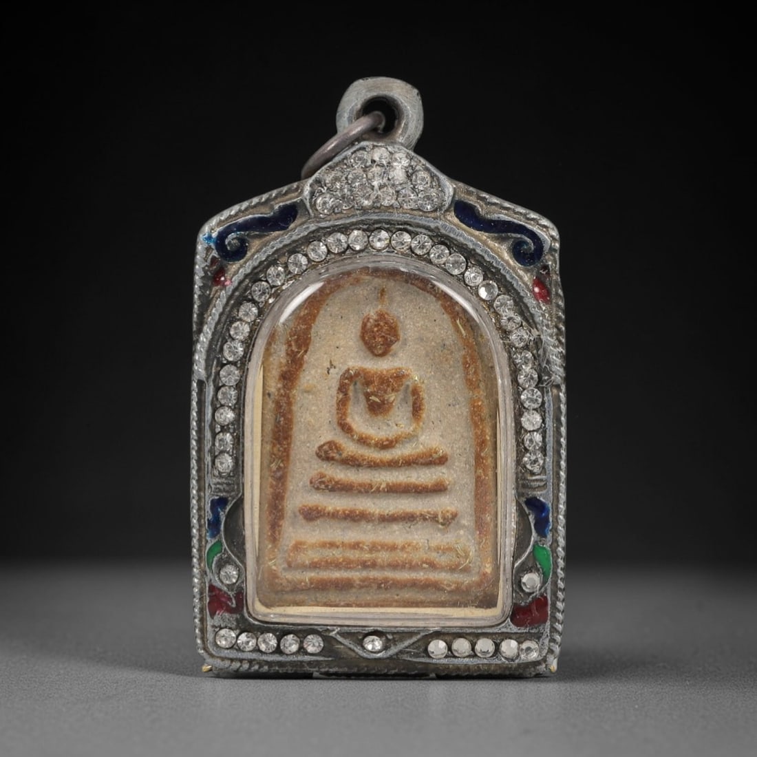 A Thai Phra Somdej Amulet in a Paste-Set Silver Frame, 20th Century, 83ZL: The amulet is a rectangular plaque with a rounded upper edge, depicting the Buddha seated in meditative pose atop a tiered base. The plaque is composed of a sacred powder composite with a pale, textur