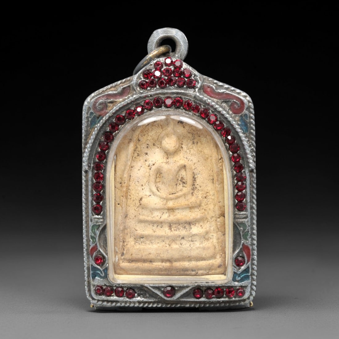 A Thai Buddhist Amulet in a Metal Case, 20th Century, 23ZK: The amulet is a clay plaque, housed in a protective metal case. The front of the case is silver-toned metal with an arched top, decorated with red gemstone inlays and enameled accents. The amulet with