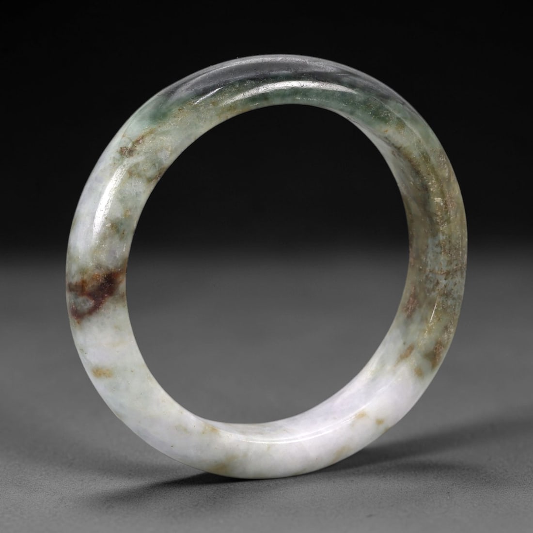 A Chinese Jadeite Jade Bangle Bracelet, 20th Century, 94ZK: Carved from jadeite jade with a variegated palette of pale lavender, greenish-grey, and irregular brownish inclusions, the bangle is formed as a continuous circular band with a rounded cross-section.