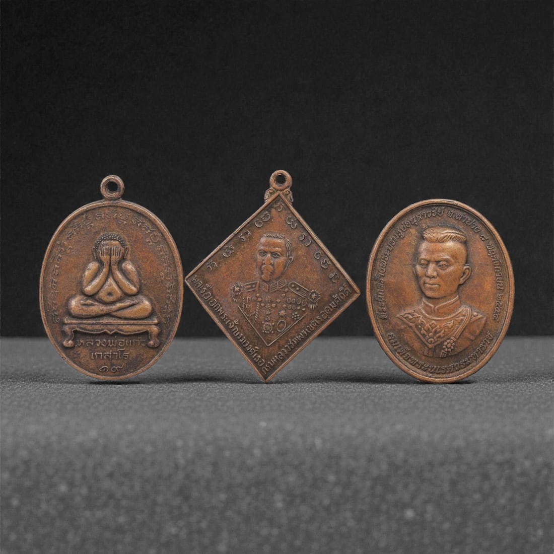 A Group of Three Thai Copper Amulets, 20th Century, 44FL: Each amulet is cast from copper with a warm brown patina, fitted with a suspension loop at the top. The first is an oval medallion depicting the Phra Pidta (closed-eye Buddha) seated on a pedestal, wi