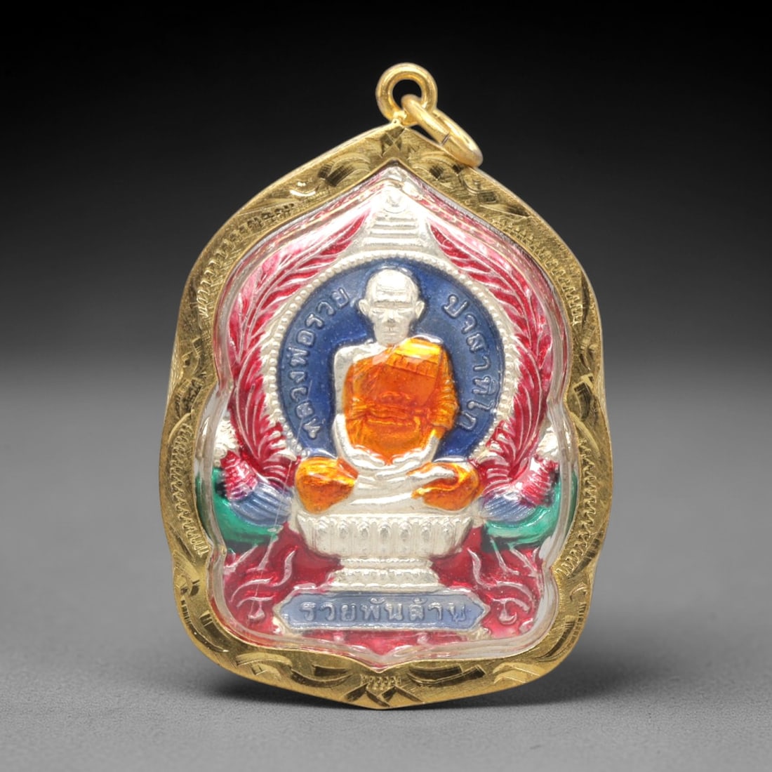A Thai Monk Commemorative Amulet in a Gilded Case, Late 20th Century, 22ZK: This ornate amulet is presented in a gilded metal case with a suspension loop at the apex. The obverse features a vibrant, multi-colored relief of a seated Thai Buddhist monk, clad in orange robes, se