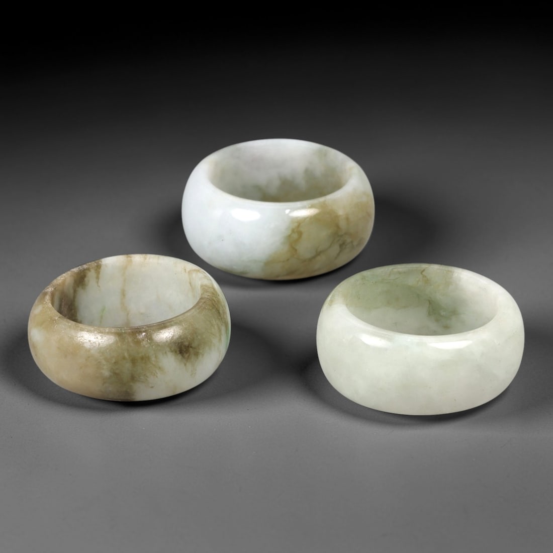 A Trio of Chinese Jadeite Rings - 20th Century, 27AL: This set of three Chinese jadeite rings features thick, rounded band forms crafted from natural jadeite with pale celadon and milky white tonalities accented by subtle mineral veining and inclusions.