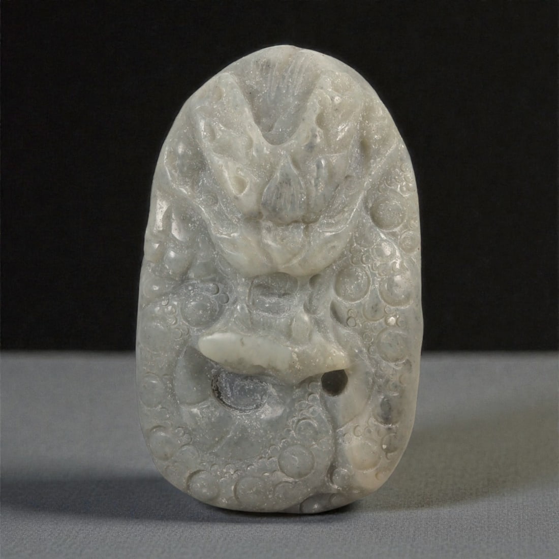 A Chinese Nephrite Jade Dragon Pendant, 20th Century, 38FB: Carved from pale grey-green nephrite jade with areas of translucency and scattered natural inclusions, the pendant is worked in an upright oval form. The surface is carved in relief with a frontal dra