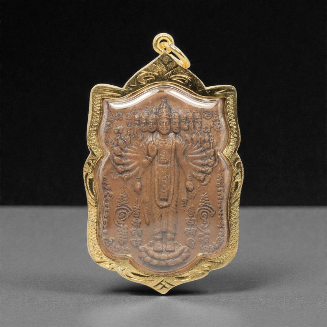 A Thai Multi-Armed Deity Amulet Pendant, 20th Century, 4FP: This shield-shaped pendant features a copper alloy core, encased in an ornate gilded metal frame with a transparent protective cover, measuring 6.2 x 3.6 cm (2 7/16 x 1 7/16 in.). The obverse displays