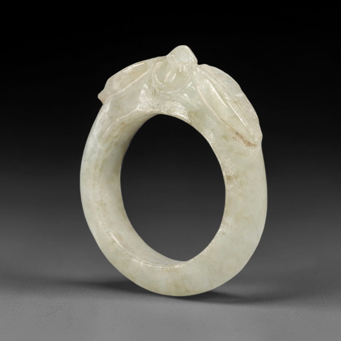 A Chinese Jadeite Jade Floral-Form Ring, Qing Dynasty, 67FS: Carved from pale greenish-white jadeite jade with natural diffuse veining and subtle internal inclusions, the object is formed as a circular ring with a raised, carved floral motif integrated into the