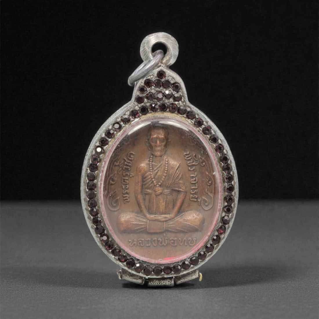 A Thai Monk Amulet Pendant with Garnet-Set Frame, 20th Century, 1FP: This oval amulet pendant features a patinated copper alloy core, encased in an ornate silver frame with a transparent protective cover, measuring 5.6 x 3.9 cm (2 3/16 x 1 9/16 in.). The obverse displa
