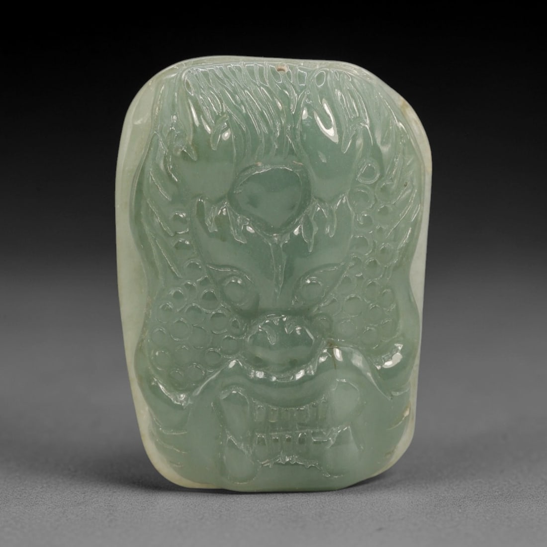 A Chinese Jadeite Jade Dragon Head Pendant, 20th Century, 19ZL: Carved from pale green jadeite with a smooth, polished surface, the object is formed as a rectangular pendant with rounded corners. The front is decorated in high relief with a stylized dragon head, f