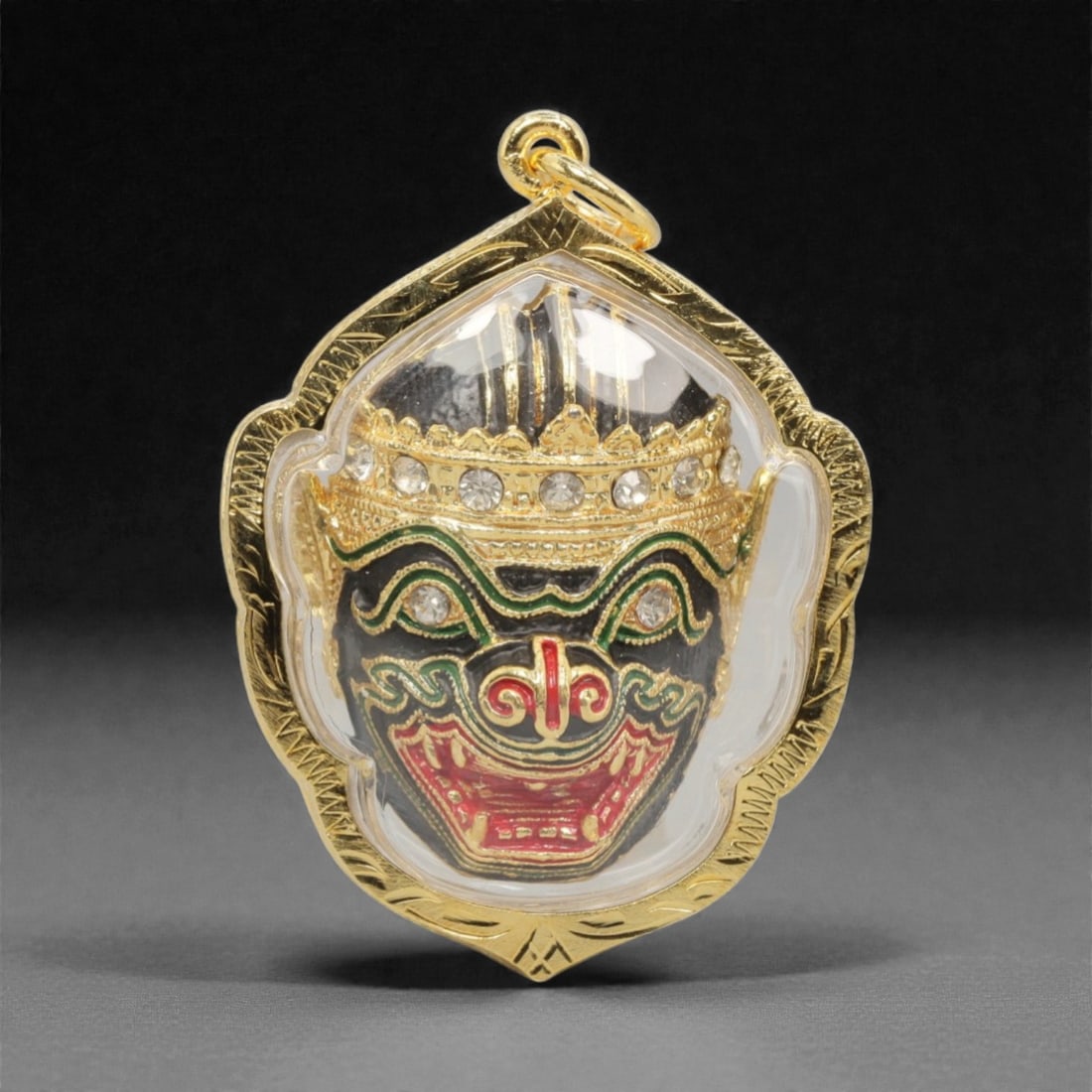 A Thai Khon Mask Amulet Pendant, 20th Century, 6FP: This pendant features a painted and gilded Khon mask at its center, set within an ornate gilded metal frame and enclosed in a transparent protective cover. The mask is rendered with a black complexion