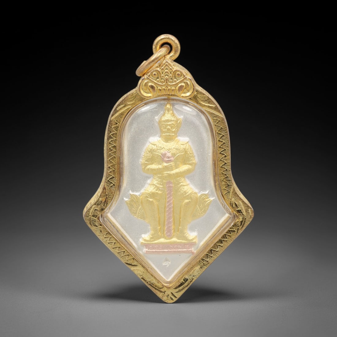 A Thai Guardian Deity Amulet in Gold Frame, Late 20th Century, 3FR: A shield-shaped amulet, measuring 5 x 3.3 cm, set within a gold frame with a suspension loop. The obverse depicts a seated guardian deity in gold and pink relief, holding a staff, enclosed in a transp