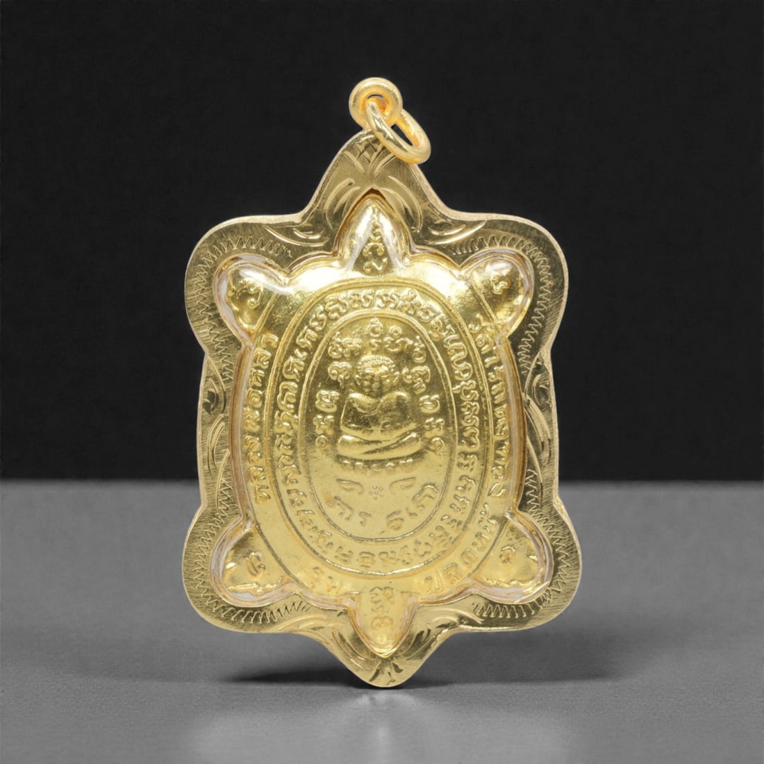 A Thai Turtle Amulet Pendant, 20th Century, 7FP: This pendant is shaped like a stylized turtle, crafted from a gilded metal and encased in a transparent protective cover. The obverse features a central seated Buddha figure, surrounded by script insc