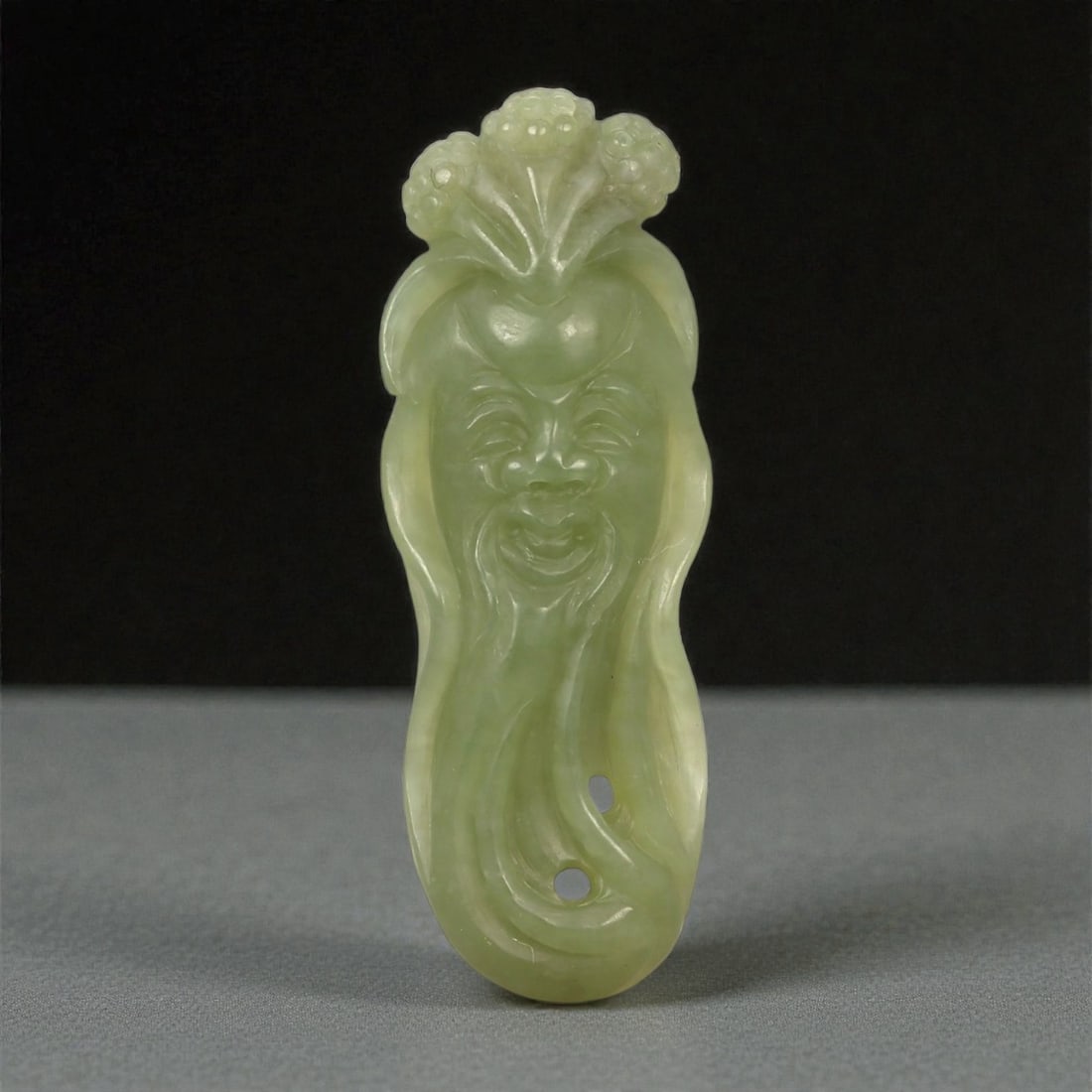 A Chinese Nephrite Jade Shoulao Pendant, 20th Century, 52FA: Carved from pale celadon nephrite jade with soft translucency and minor natural inclusions, the pendant depicts Shoulao, the Daoist god of longevity, shown frontally with an elongated face and flowing