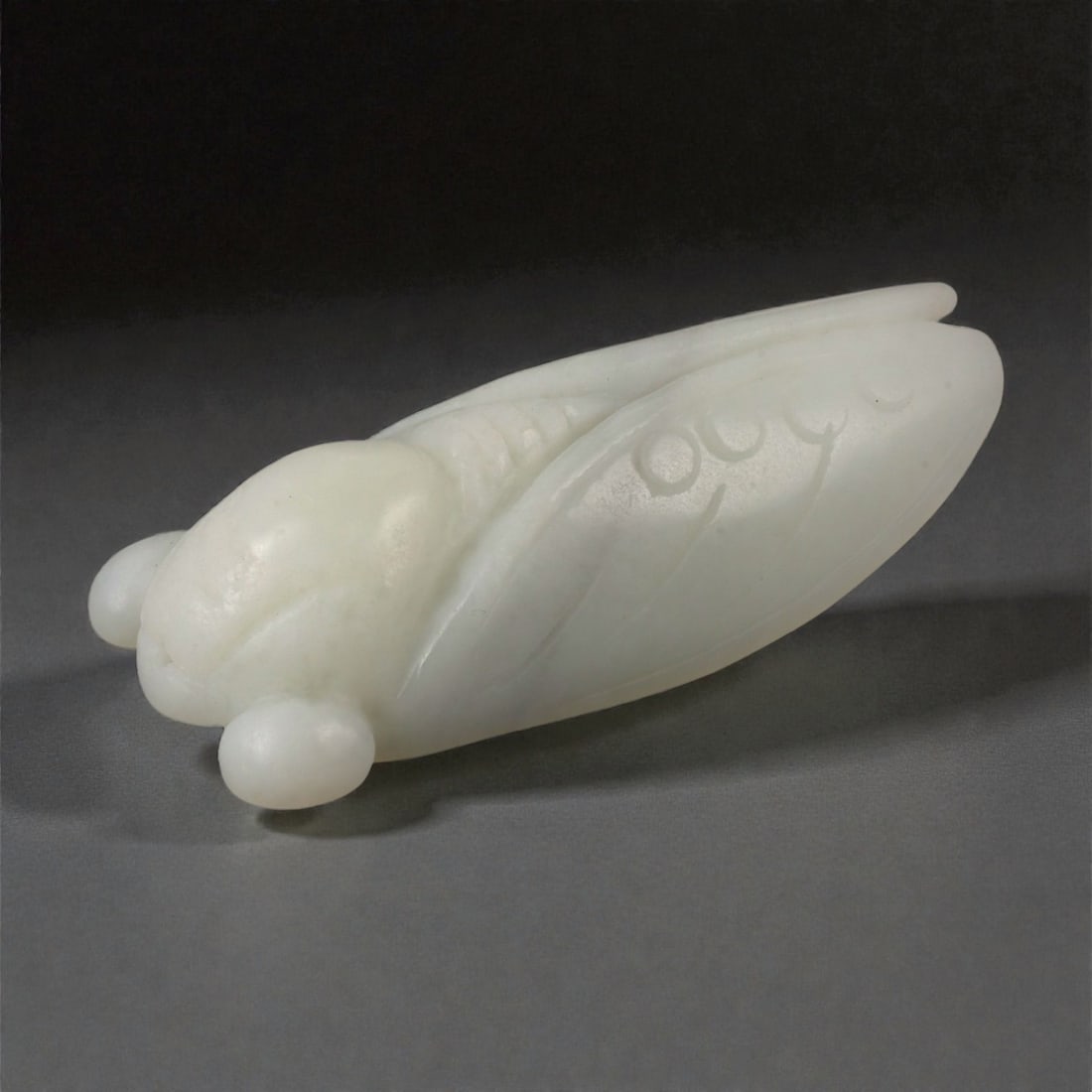 A Chinese Nephrite Jade Cicada Amulet, 20th Century, 80FB: Carved from pale celadon Hetian nephrite jade with even translucency and a softly polished surface, the amulet is formed as a cicada shown in simplified naturalistic relief. The body is elongated and