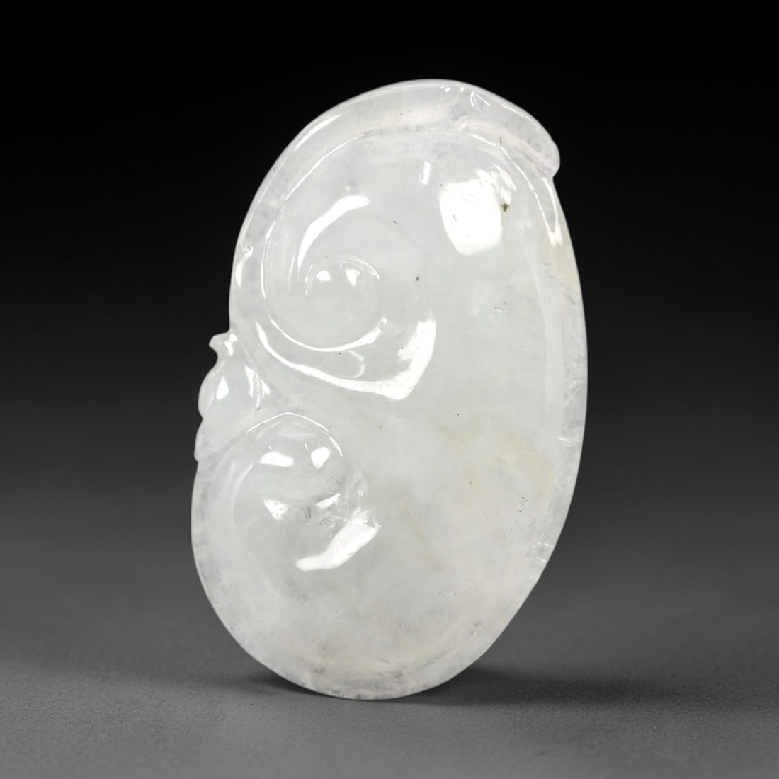 A Chinese Icy Jadeite Jade Ruyi Pendant, 20th Century, 55AB: Carved from translucent icy jadeite with a pale milky-white hue and minor natural inclusions, the pendant is shaped as a stylized ruyi scepter form. Two raised, spiraled scroll motifs are carved in lo