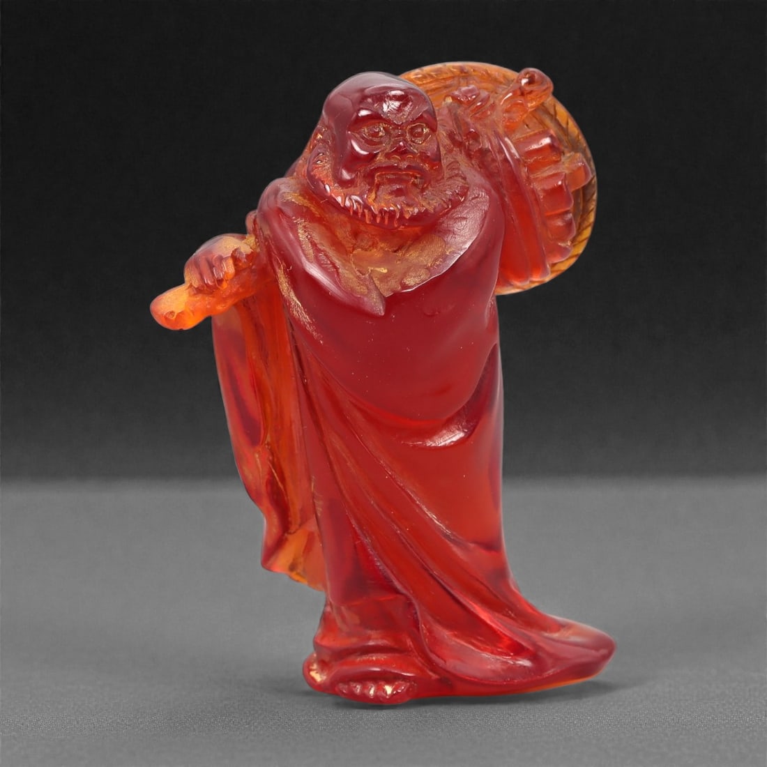 A Chinese Amber-Resin Figure of a Luohan, 20th Century, 41FH: Cast or carved from translucent amber-colored red resin with internal tonal variation, scattered inclusions, and minor entrapped bubbles, the figure represents a standing luohan shown holding a staff